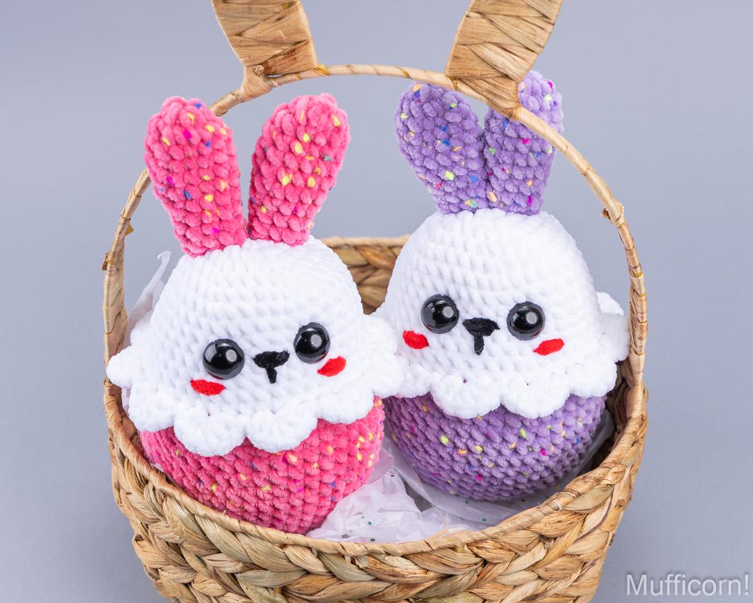 Two crocheted bunny-shaped plush toys sitting in a woven straw basket. One bunny is white with pink ears and pink body, and the other is white with purple ears and purple body.