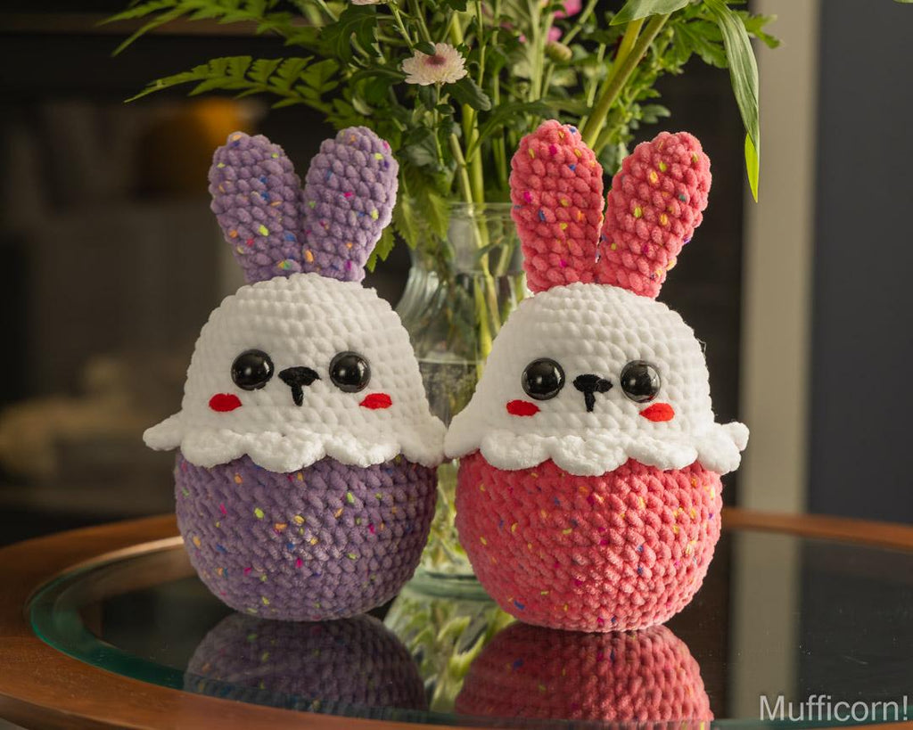 Two handmade crocheted bunny sitting on a glass table in front of a vase of flowers. One doll is purple and white, and the other is pink and white, both with embroidered black eyes and red cheeks.
