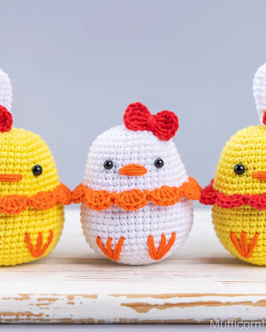Three crocheted Easter ducks on a rustic white wooden surface against a soft gray background. The center figure is white with an orange beak, orange feet, and a red bow, flanked by two yellow chicks with red and orange accents.