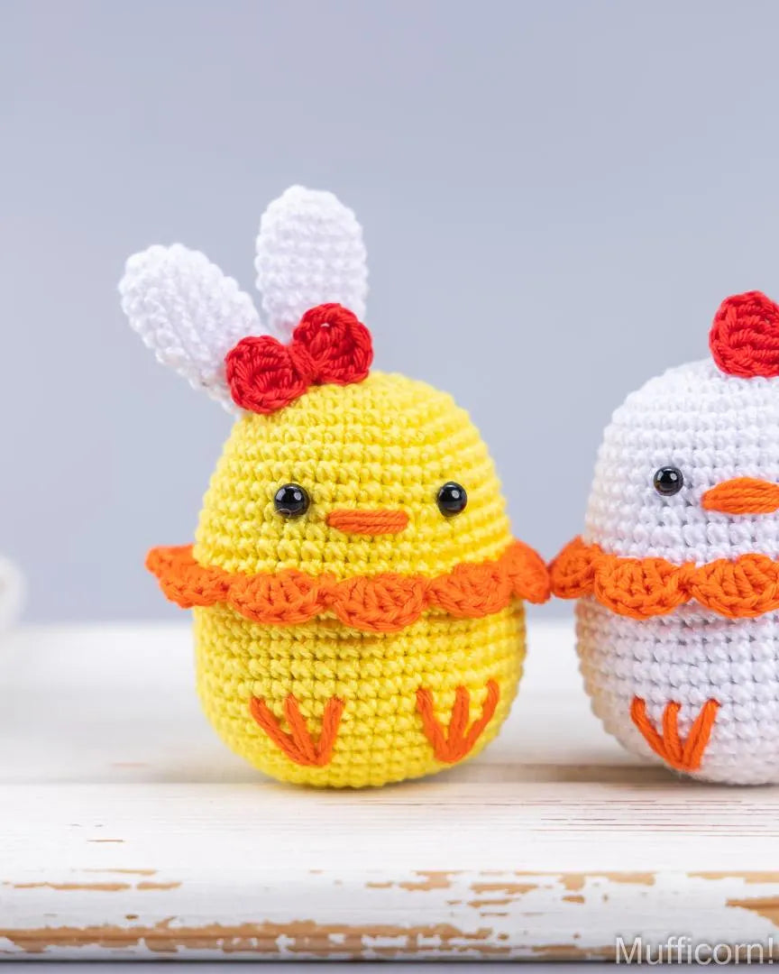 Two crocheted Easter duck on a rustic white wooden surface against a soft gray background. The left chick is yellow with white bunny ears, a red comb, and an orange ruffled collar.