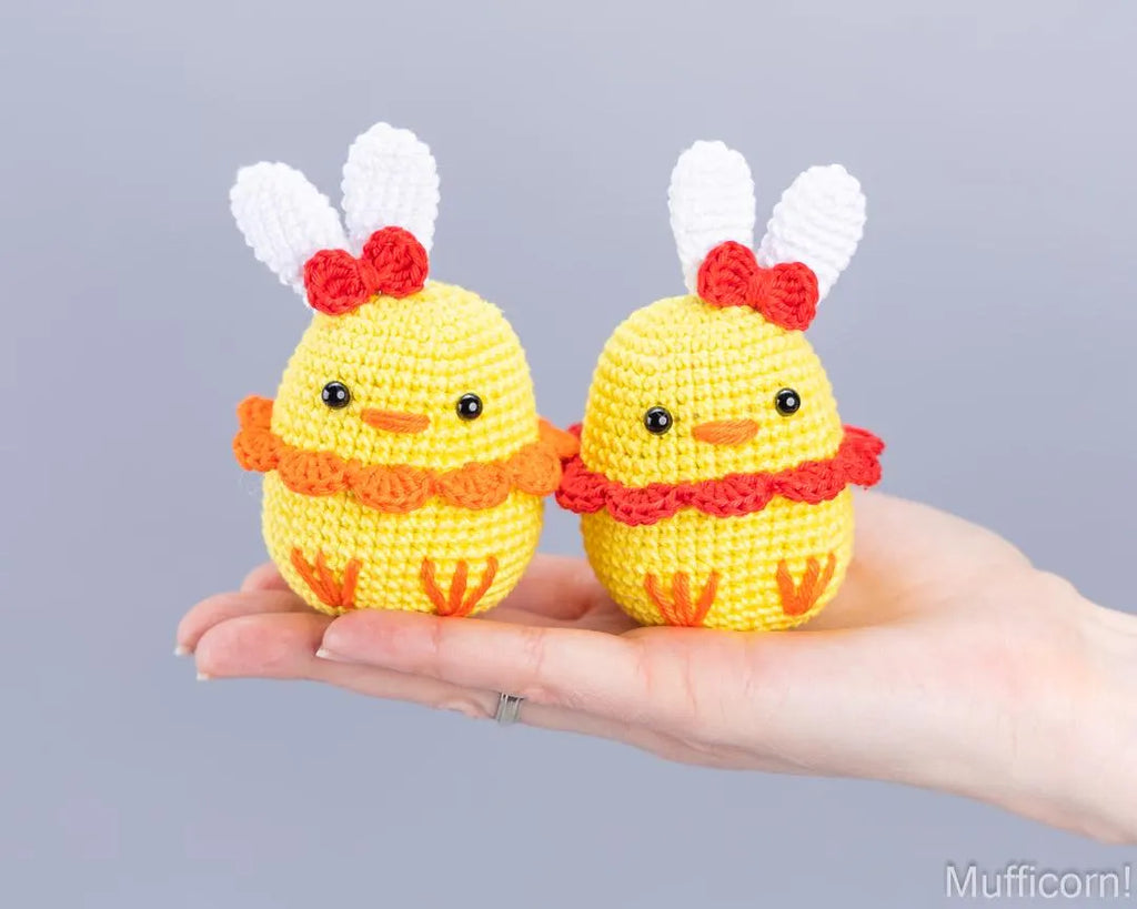 Two small crocheted yellow duck toys with white bunny ears, red bows, and orange accents are held in a person's hand against a plain light gray background.