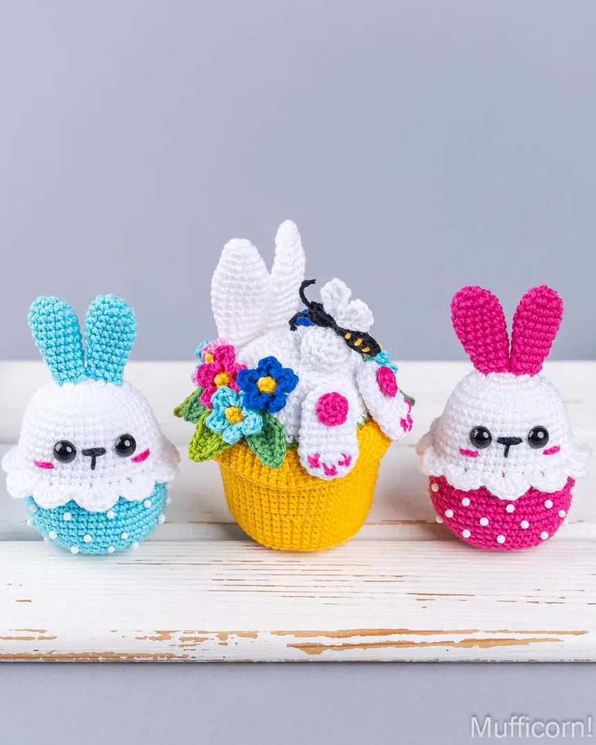 Three crocheted bunny characters arranged on a white rustic surface against a light gray background. The left bunny is white with turquoise ears and a turquoise dotted bottom.
