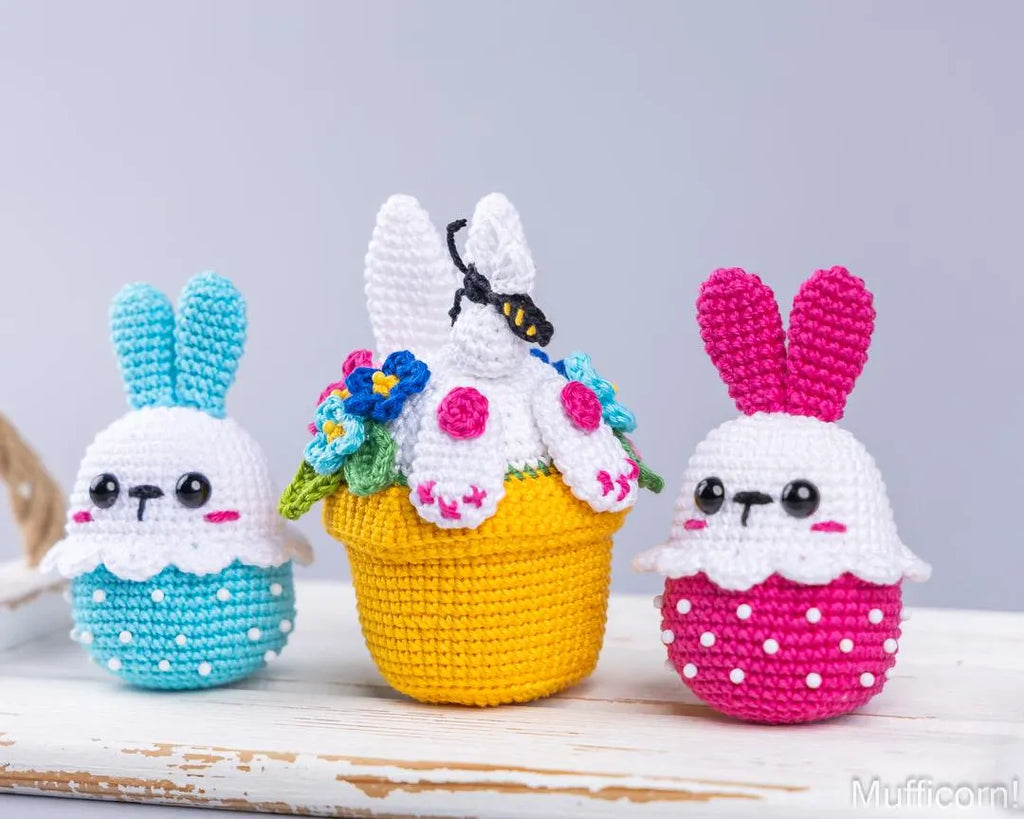 Three crocheted amigurumi Easter-themed characters on a rustic white wooden surface against a light gray background: a blue bunny on the left, a yellow cupcake with white frosting and a black-and-yellow bee in the center, and a pink bunny on the right.