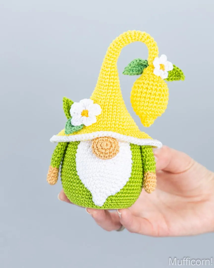 A crocheted gnome doll with a yellow hat shaped like a lemon, green body, white beard, and small flower details, being held in a person's hand against a light gray background.