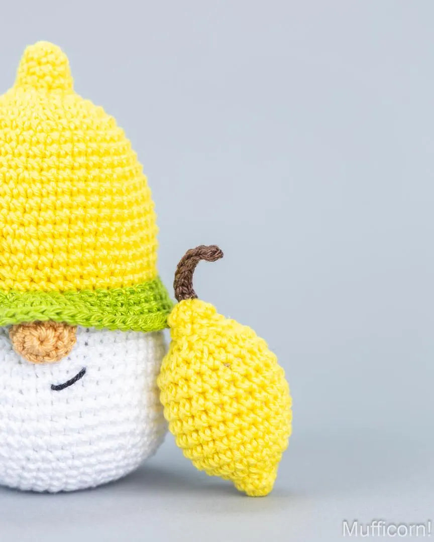 A crocheted amigurumi character resembling a lemon gnome, featuring a yellow crocheted hat with a green band, a white body, a small brown nose, and a yellow lemon-shaped accessory with a brown stem, photographed on a light gray background.