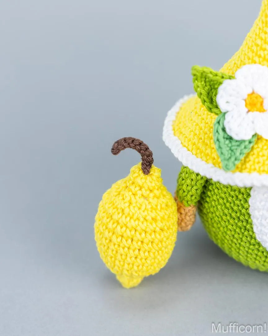 Close-up of a crocheted yellow lemon character with a brown stem next to a crocheted character wearing a yellow and green outfit with a white flower, on a light gray background.