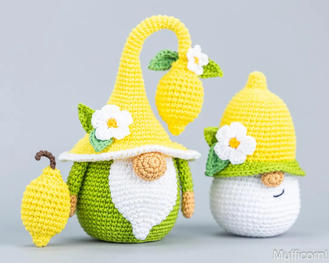 Two crocheted lemon-themed gnomes on a light gray background. One gnome wears a tall yellow hat with a hanging lemon and a white flower, while the other wears a shorter yellow hat with a green band and white flower.