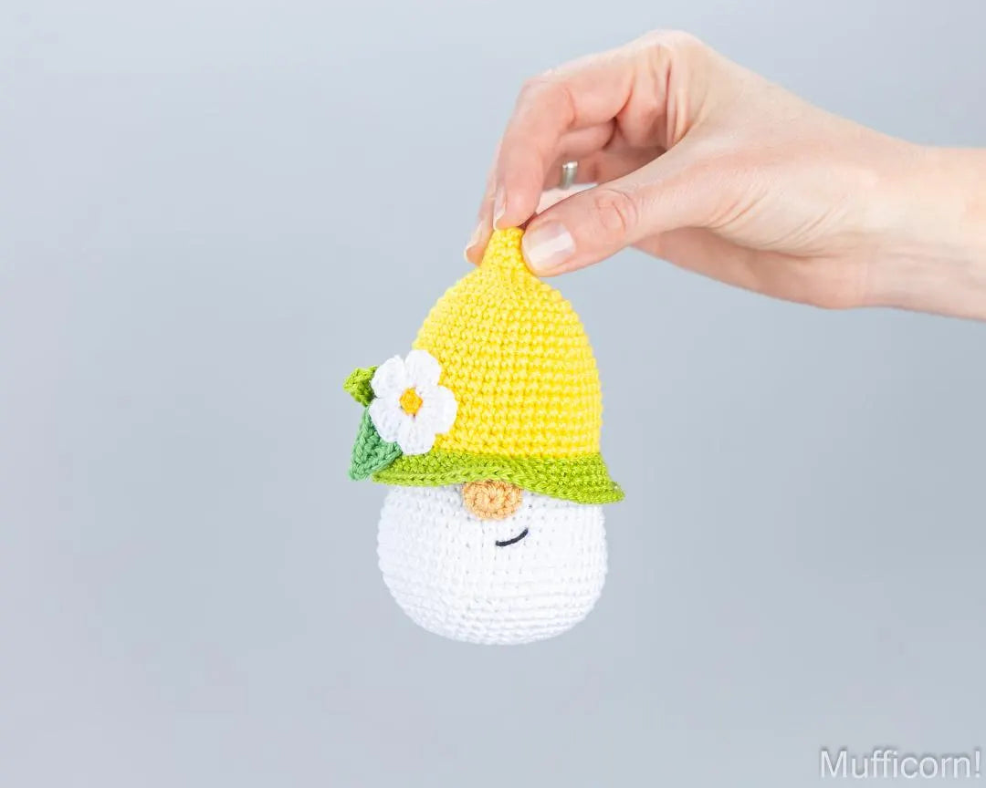 A hand holds a crocheted muffin shaped with a yellow and green hat decorated with a white flower, against a plain light gray background.