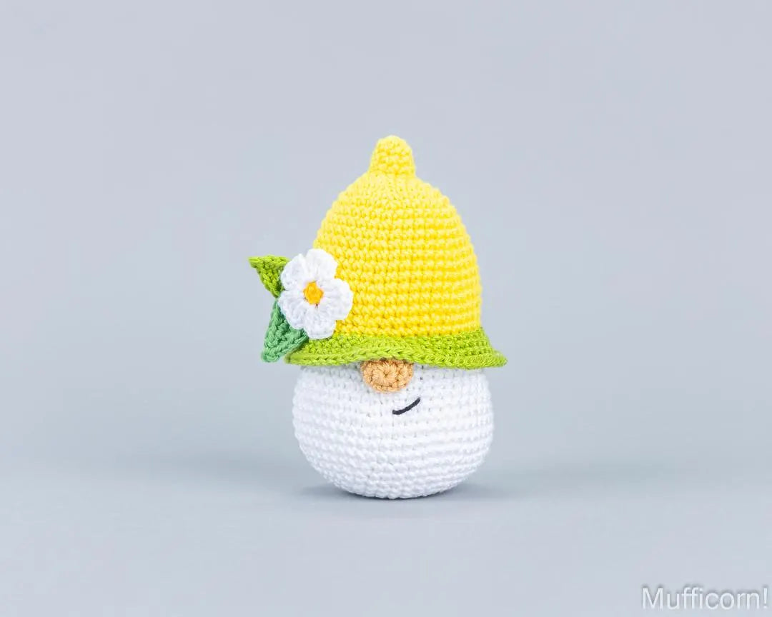 A crocheted lemon gnome bag charm with a white body, a yellow hat, and a green leafy brim decorated with a white flower, set against a solid light gray background.