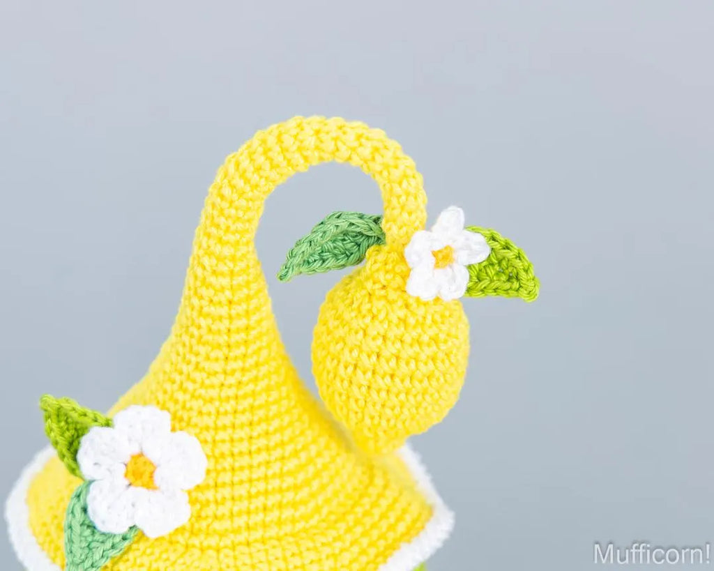 A close-up of a crocheted yellow lemon-shaped accessory with green leaves and white daisy details, photographed against a soft light gray background.