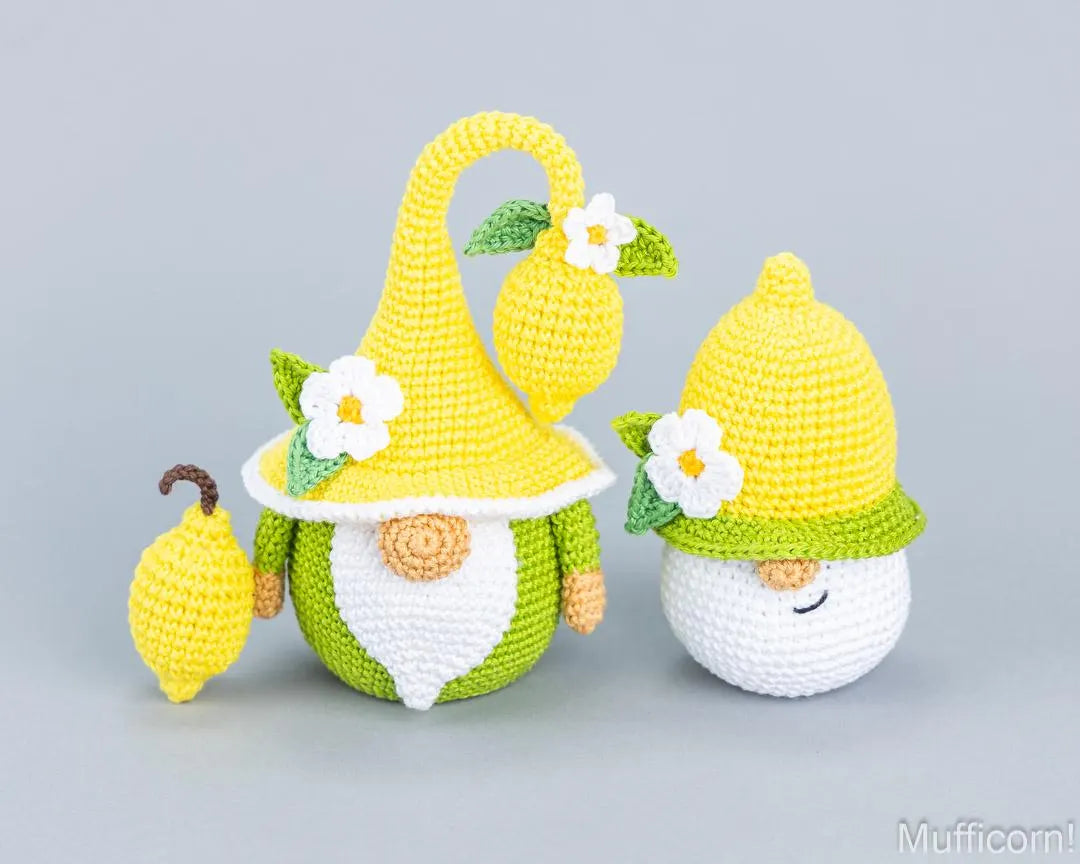 Two crocheted gnome figures and a small hanging lemon decoration in bright yellow and green, shown on a plain light gray studio background. One gnome is fully visible with a white face and closed smile, and the other is partially hidden behind the first.