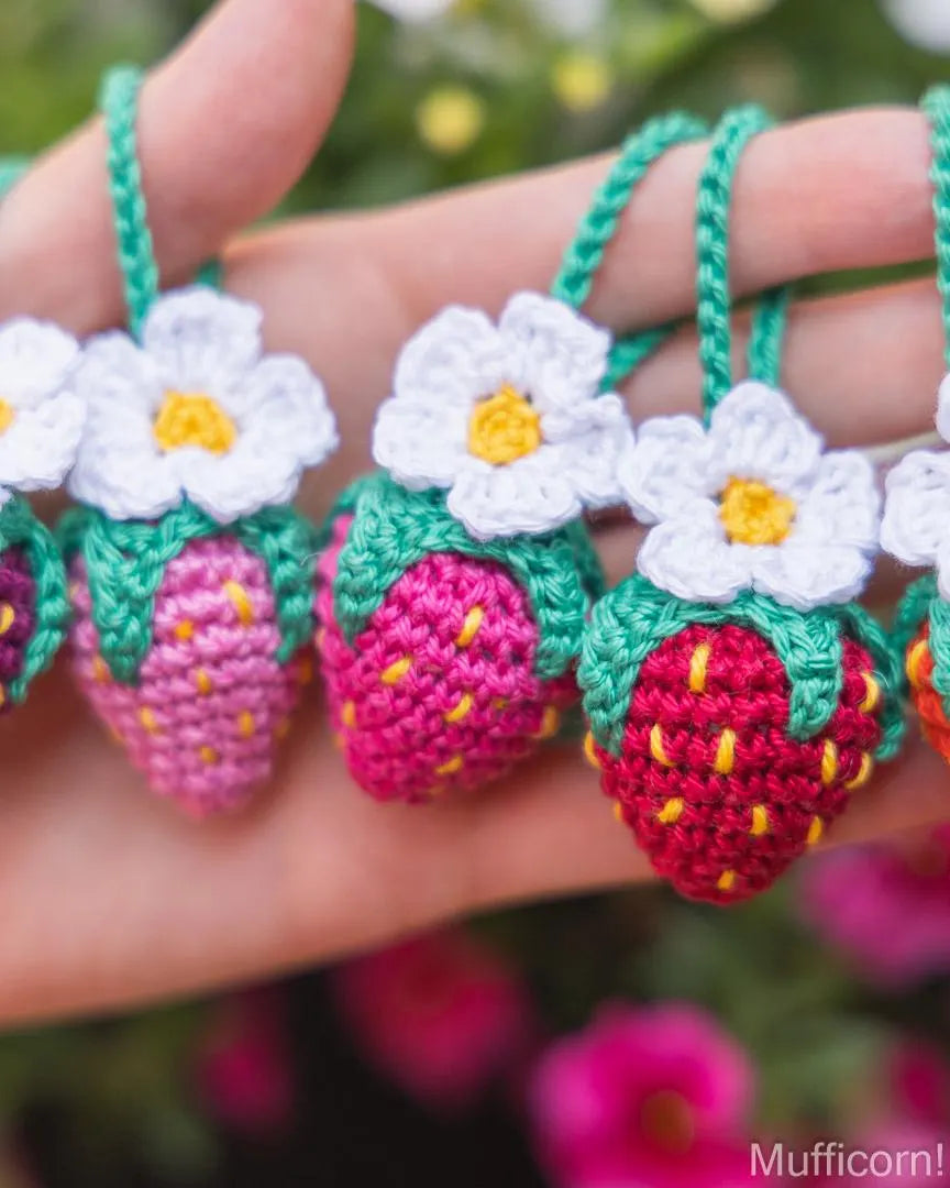 A close-up of a hand holding several crocheted strawberry charms with white flowers and green hanging loops, against a blurred garden background.