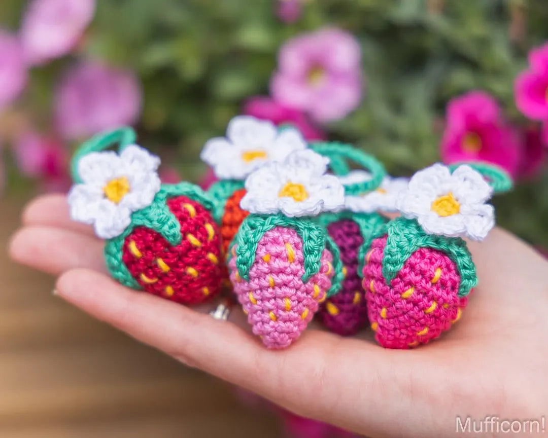 Three crocheted strawberry keychains with white daisy flowers are held in a person's hand against a soft-focus background of pink flowers and green foliage.