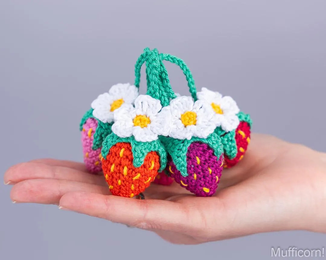 A hand holding a small crocheted strawberry-shaped purse with white daisy flowers and green handles, shown against a plain light gray background.