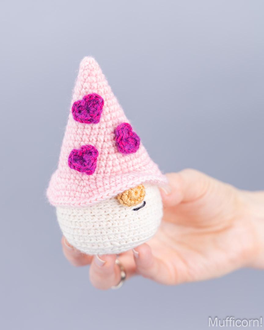 A hand holding a small crocheted gnome doll with a pink pointed hat decorated with three pink hearts, a white body, and a small tan nose, against a plain light gray background.