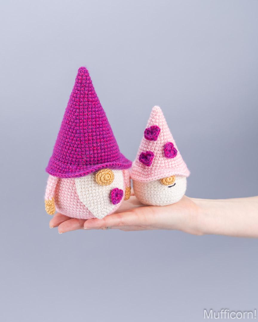 Two crocheted gnome dolls are held in a person's hand against a plain light gray background. One gnome has a bright pink hat and white body, while the other has a pale pink hat with purple flowers and a cream body.