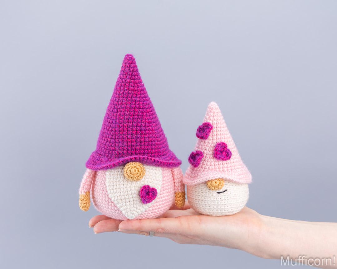 Two crocheted gnome dolls are held in a person's hand against a plain light gray background. The larger gnome wears a bright pink hat and has a pink-and-white striped body, while the smaller gnome wears a pale pink hat with pink flowers and a cream body.