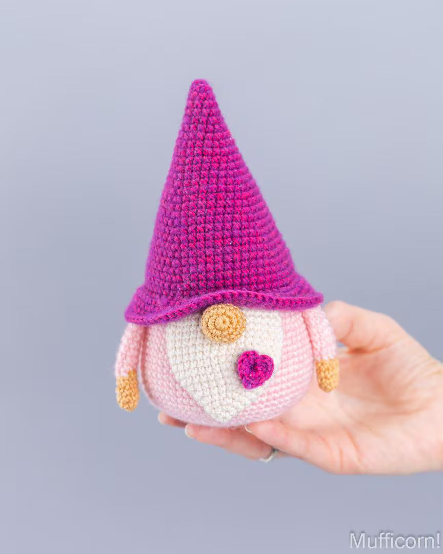 A crocheted gnome doll with a bright pink hat, pale pink body, and cream face is being held in a person's hand against a plain light gray background.