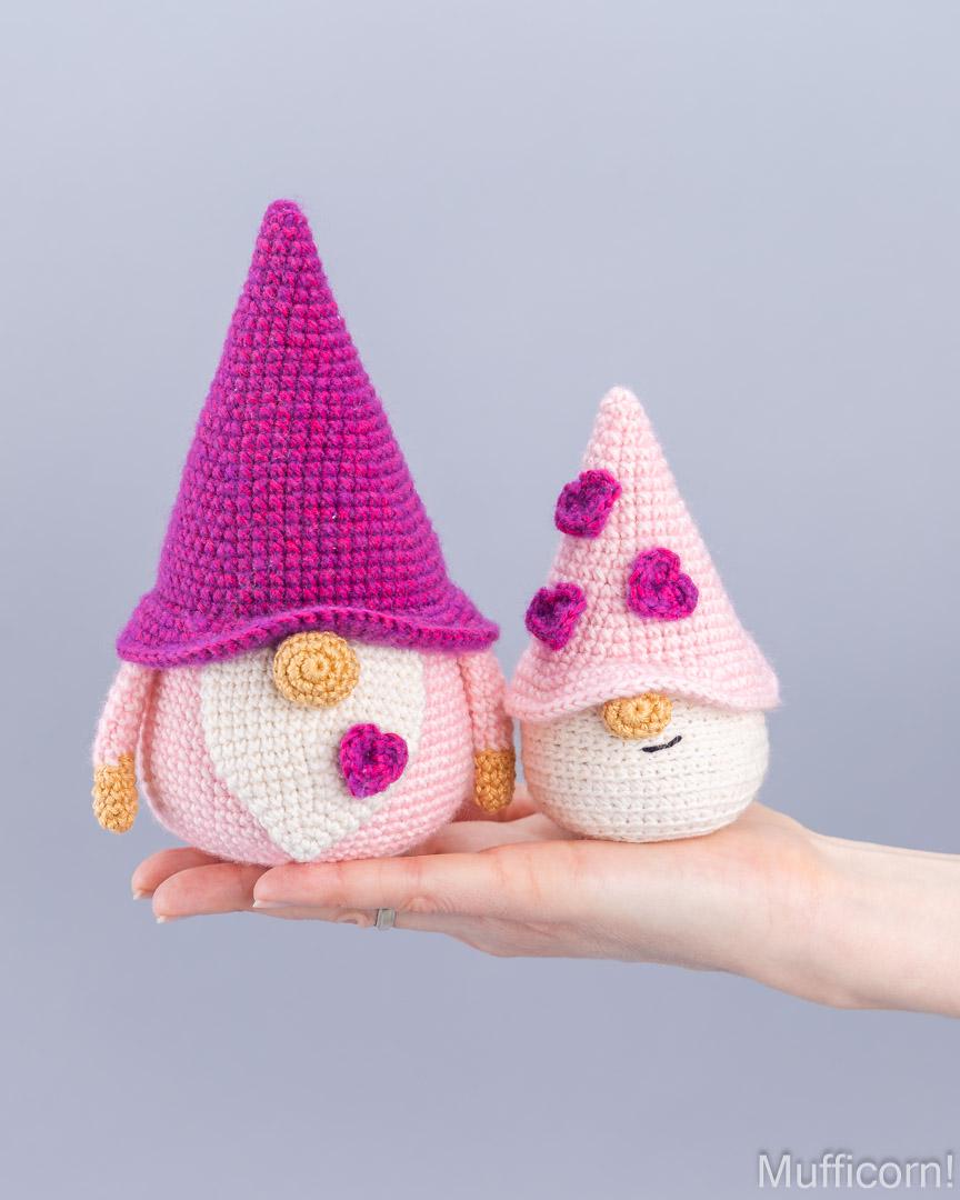 Two crocheted gnome dolls are displayed on a person's hand against a solid light gray background. The larger gnome has a bright magenta hat, a white face, and a pink body.