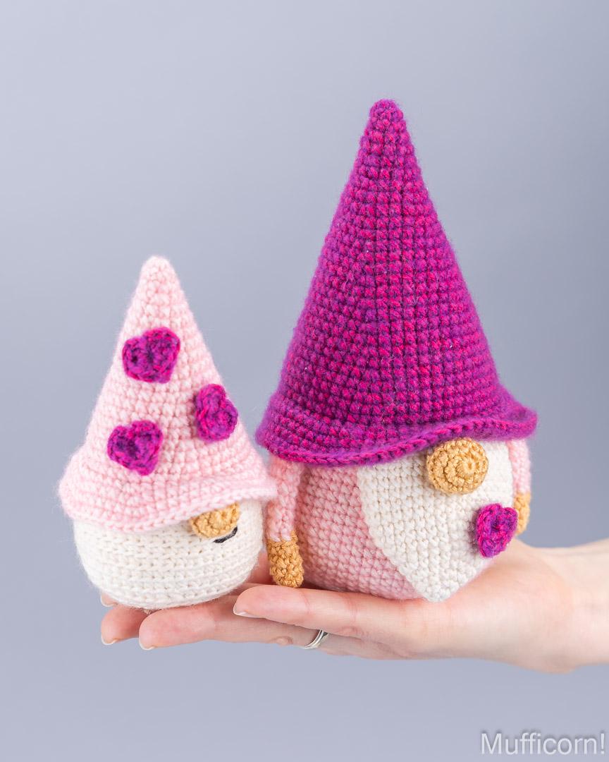 Two crocheted amigurumi garden gnomes held in a person's hand against a plain light gray background. One gnome has a pink hat with purple hearts and cream body; the other has a bright pink-purple hat, cream body, and gold details.