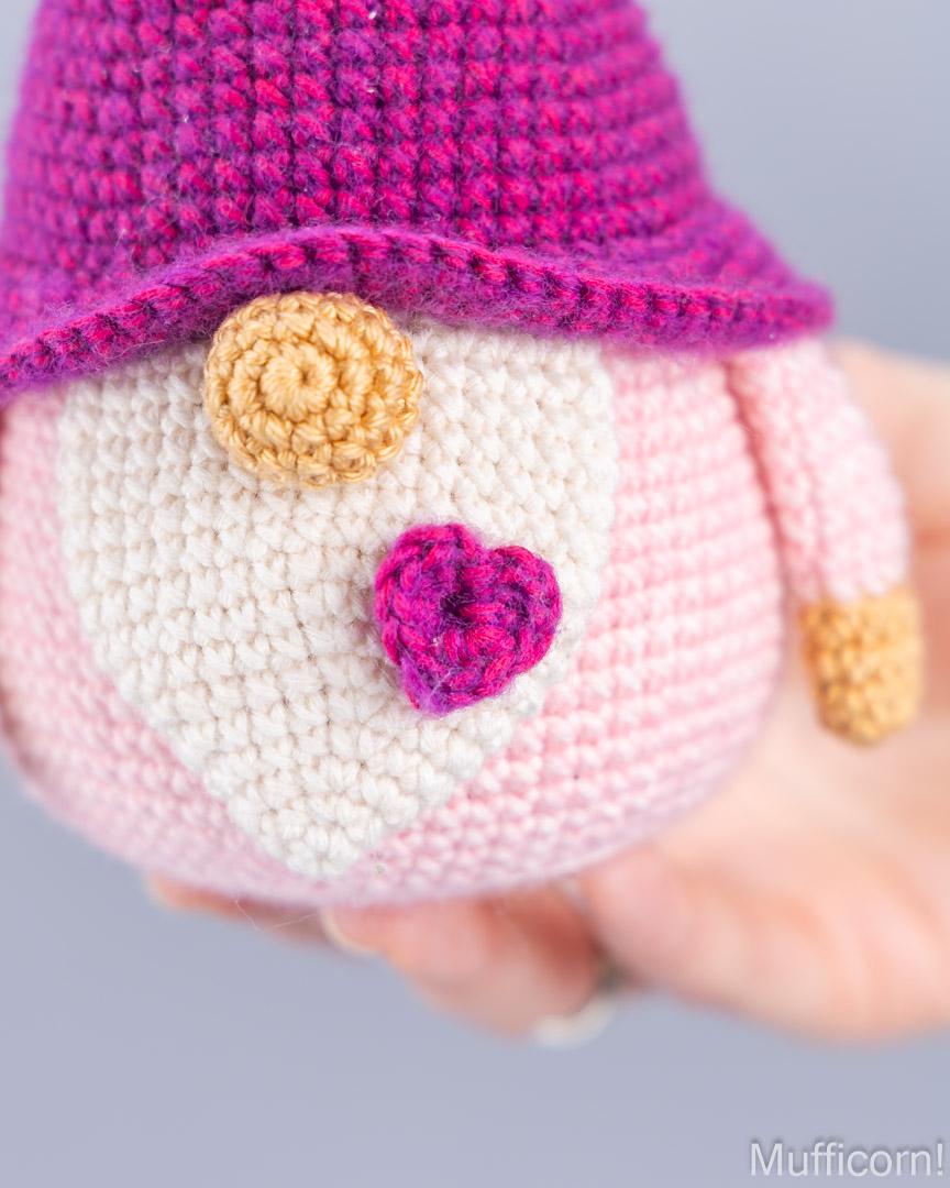 A close-up of a crocheted plush toy being held in a hand. The toy has a pink body, a purple hat, a cream-colored face, and small decorative facial details.