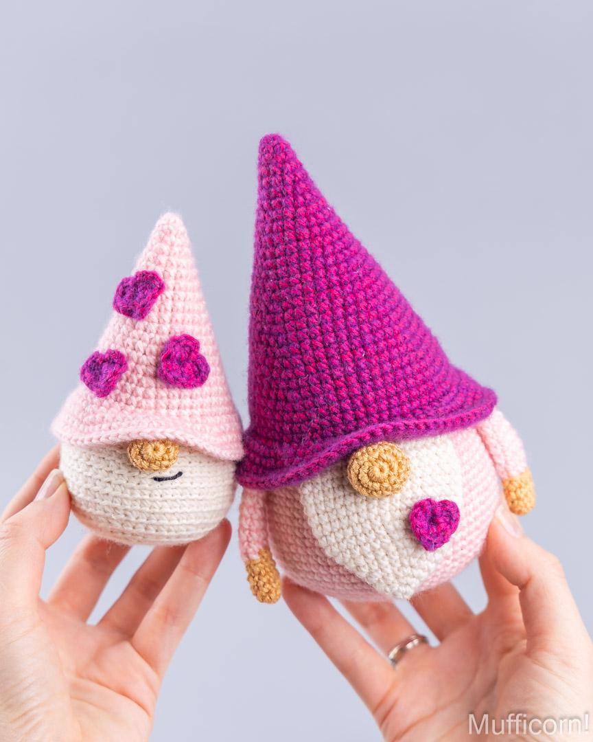 Two crocheted gnome dolls are held in hands against a light gray background. One gnome has a pink hat with purple hearts and a cream face, while the other has a magenta and purple pointed hat, a cream face, and small gold details.