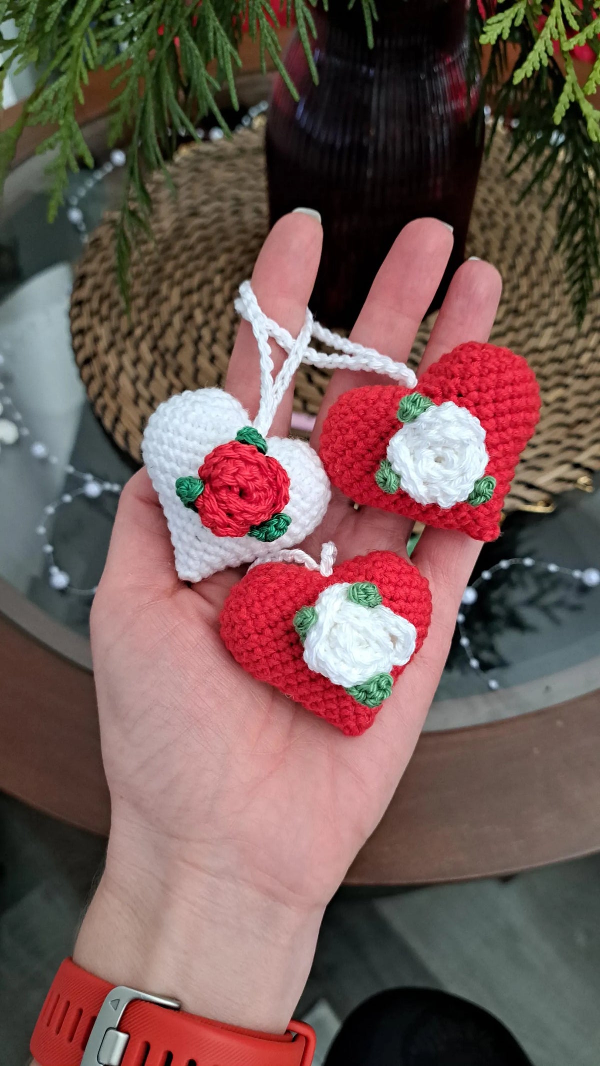 Three small crocheted heart ornaments with red and white roses are held in a person's hand. The ornaments have white hanging loops and are decorated with red and white yarn roses with green leaves.