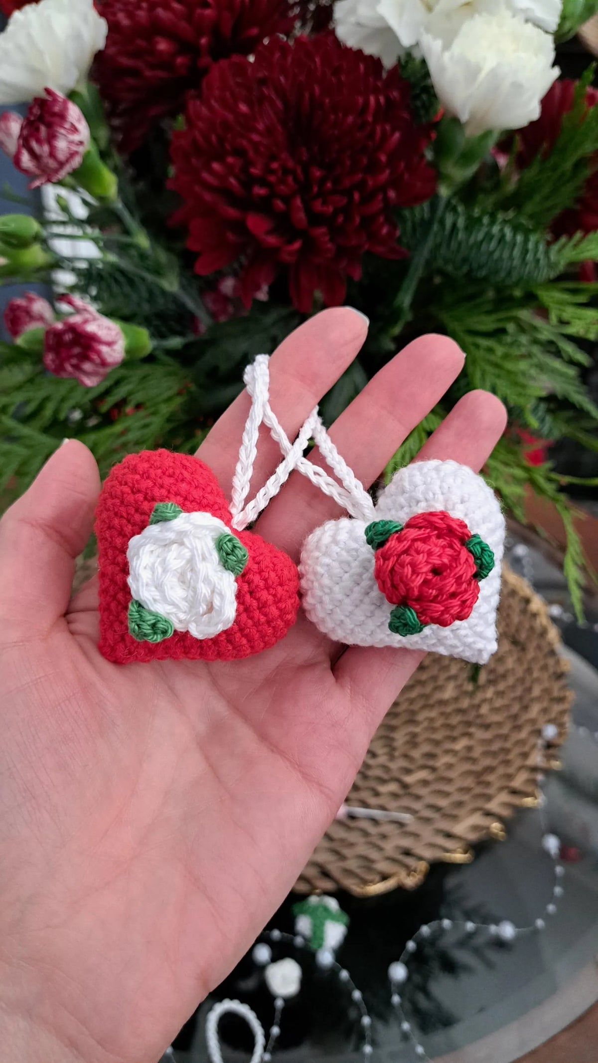 A hand holds two small crocheted heart ornaments in front of a bouquet of red and white flowers. One heart is red with a white rose and green leaves, and the other is white with a red rose and green leaves.
