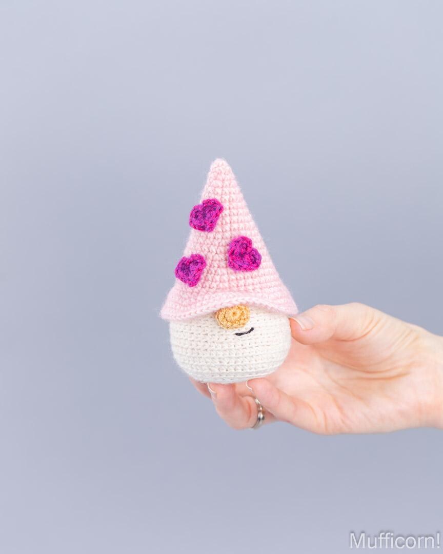 A hand holding a small crocheted gnome doll with a pink pointed hat decorated with three purple flowers, a white body, and a small yellow nose, against a plain light gray background.
