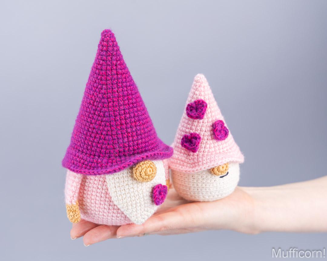 Two crocheted garden gnomes displayed on a person's hand against a soft gray studio background. The larger gnome wears a bright magenta-pink pointed hat and a cream-colored beard, while the smaller gnome wears a pale pink hat with purple flower details and a cream face.