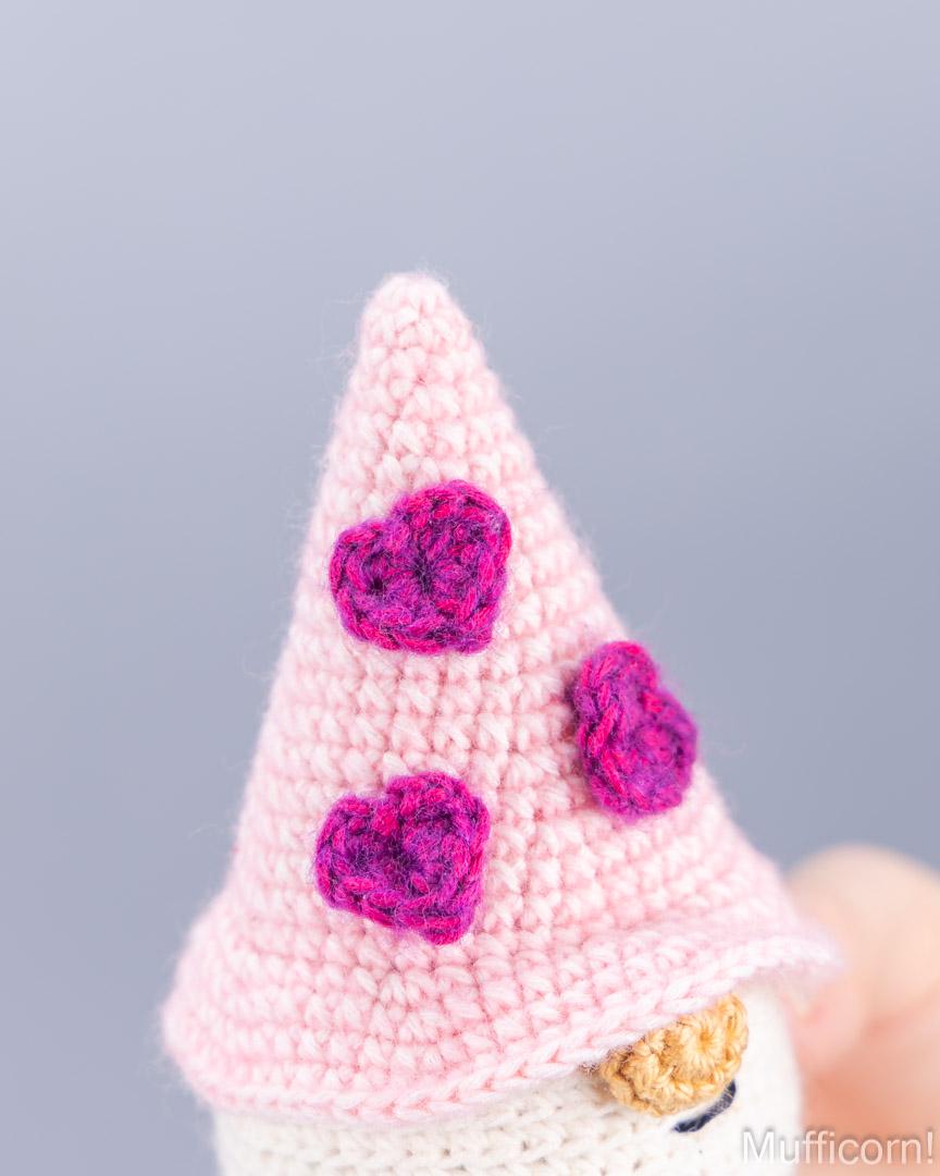 Close-up of a crocheted gnome hat in light pink with three bright magenta heart accents, shown against a soft gray background.