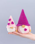 Two crocheted gnome dolls are held in a person's hand against a plain light gray background. The larger doll wears a bright pink hat and has a pink heart detail, while the smaller doll wears a pale pink hat with pink flowers.