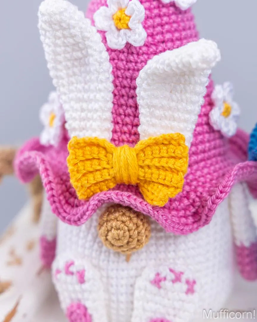 Close-up of a crocheted bunny character wearing a pink hat with white ears, white flowers, and a yellow bow, with a white body and pink details.