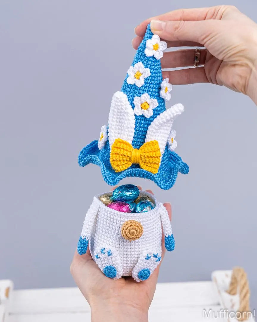 A crocheted Easter-themed pot holder and lid set, held by hands against a soft gray background. The white crocheted pot is filled with colorful chocolate eggs, and the matching blue, white, and yellow crocheted lid is being lifted by a hand.