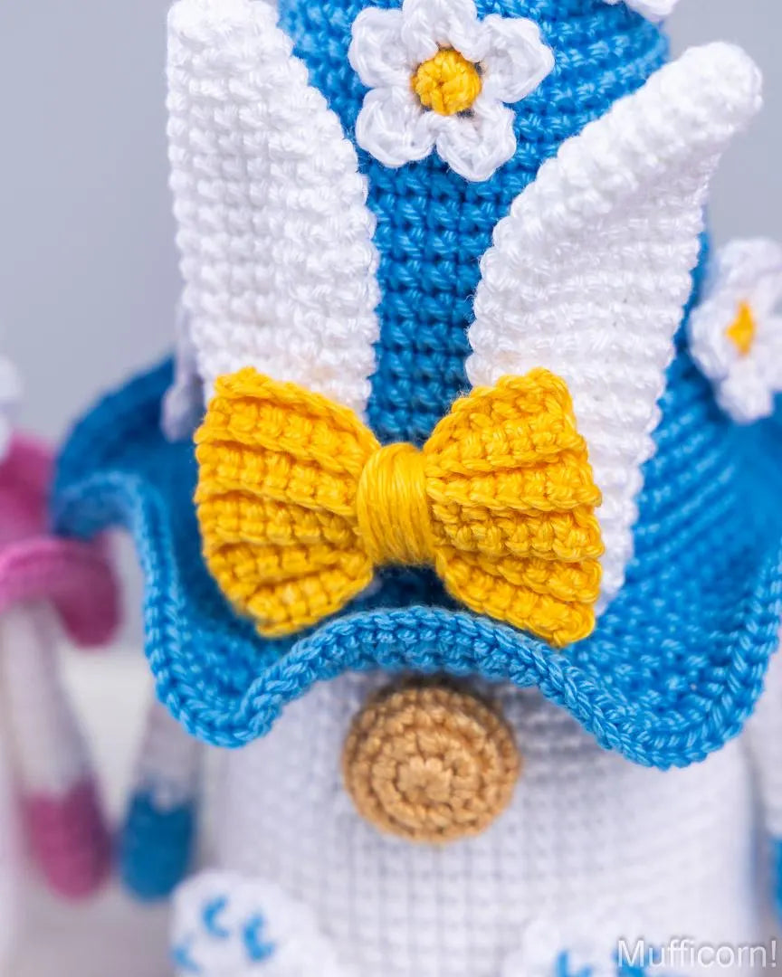 Close-up of a crocheted character hat with white wings, a bright blue brim, a yellow crocheted bow, and white flower accents on a light gray background.