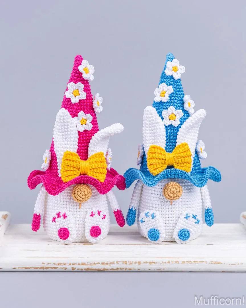 Two crocheted bunny gnomes with pointed hats and yellow bows, displayed side by side on a white wooden surface against a light gray background.