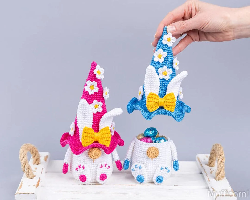 Three crocheted Easter gnomes displayed on a white wooden surface against a light gray background. A human hand is holding one blue gnome above the others.