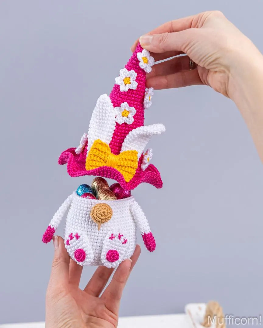 A crocheted Easter gnomemuffler being held up by two hands. The white and pink crocheted gnomemuffler has a yellow bow, white daisy accents, and a pointed hat, with a small basket filled with chocolate eggs tucked underneath.
