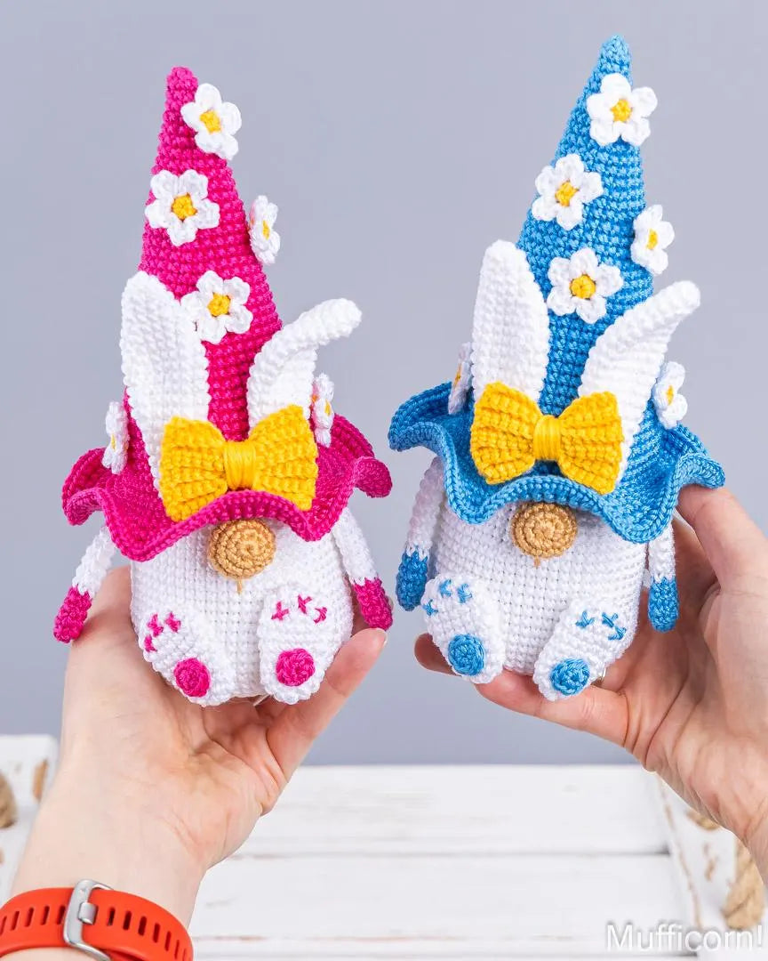 Two crocheted bunny gnomes held in hands against a soft gray background. One gnome has a pink hat and collar with white daisy flowers and a yellow bow, and the other has a blue hat and collar with white daisy flowers and a yellow bow.