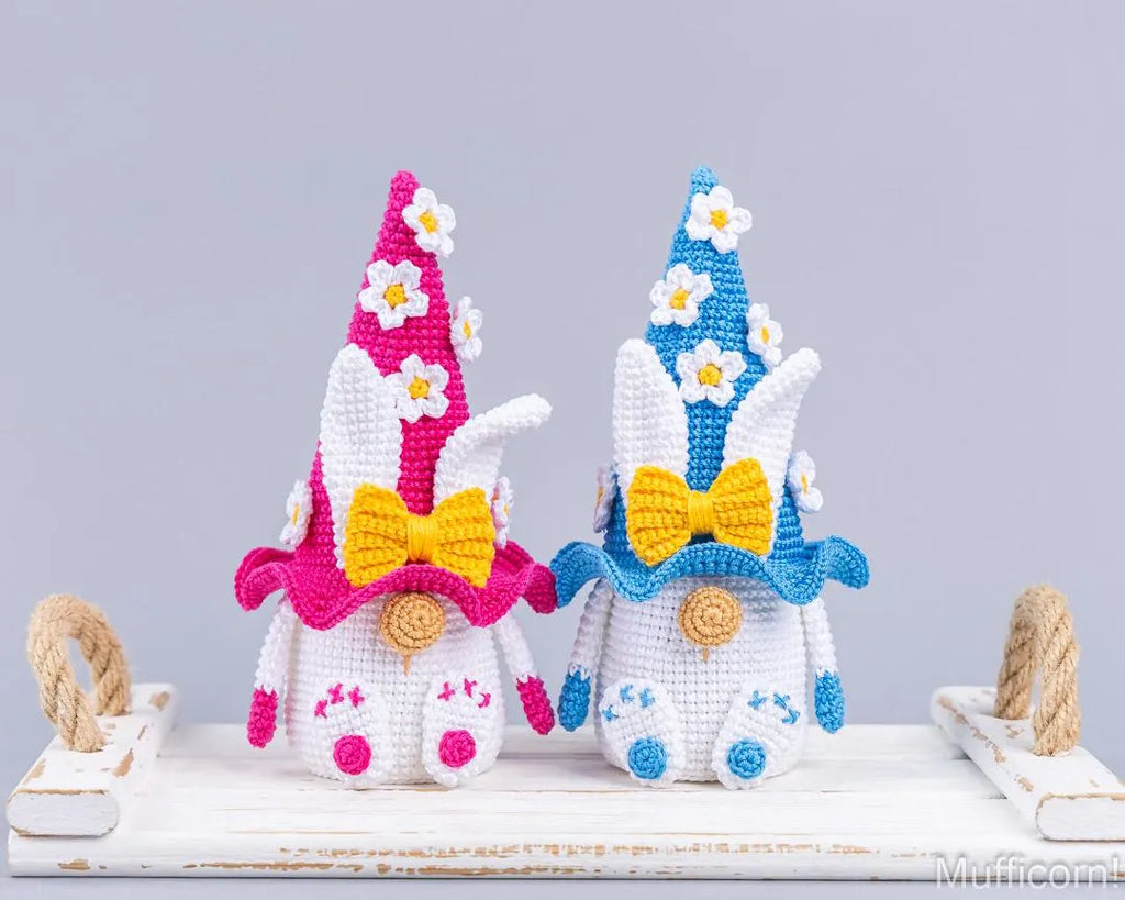 Two crocheted bunny garden gnomes displayed side by side on a white wooden surface. The left gnome has a pink hat with white daisy flowers, a yellow bow, and blue accents.