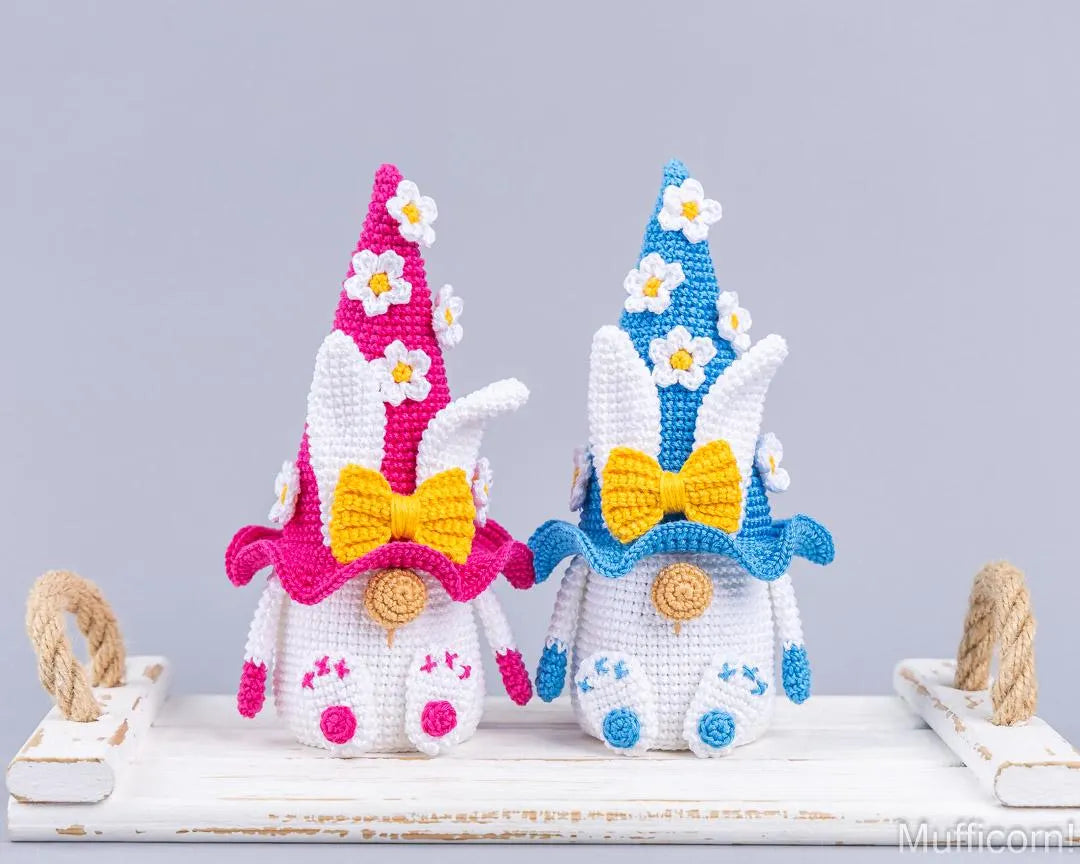 Two crocheted bunny garden gnomes displayed side by side on a white wooden surface. The left gnome has a pink hat with white daisy flowers, a yellow bow, and blue accents.