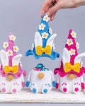 Three crocheted Easter bunny ornaments wearing pointed hats are displayed on a white surface against a light gray background. A human hand is holding the blue hat in the center.