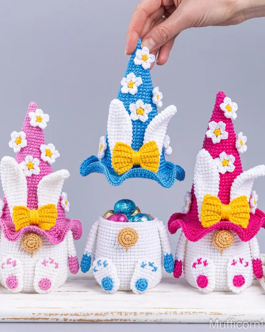 Three crocheted Easter bunny ornaments wearing pointed hats are displayed on a white surface against a light gray background. A human hand is holding the blue hat in the center.