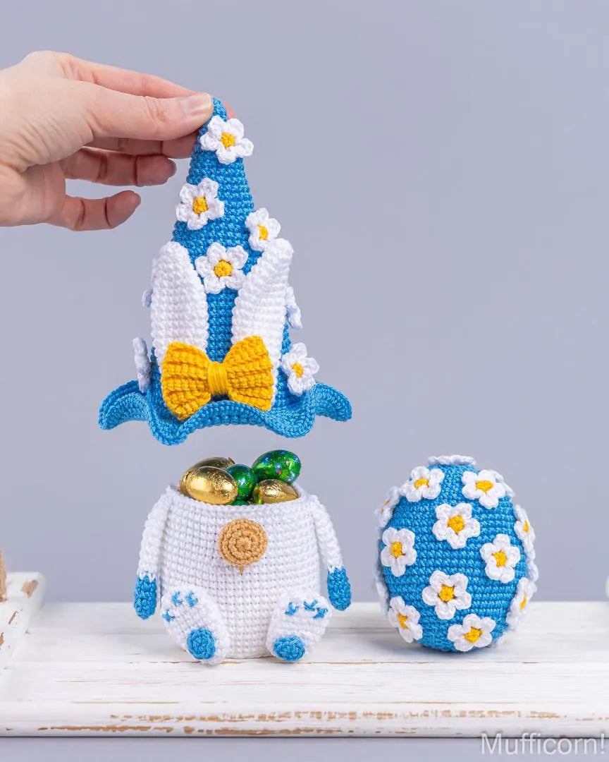 A crocheted Easter-themed pot and lid set with a blue and white pointed hat lid decorated with white daisy flowers and a yellow bow, shown being held by a hand. The pot is white with blue accents and contains chocolate coins, next to a matching blue and white crocheted egg on a white wooden surface.