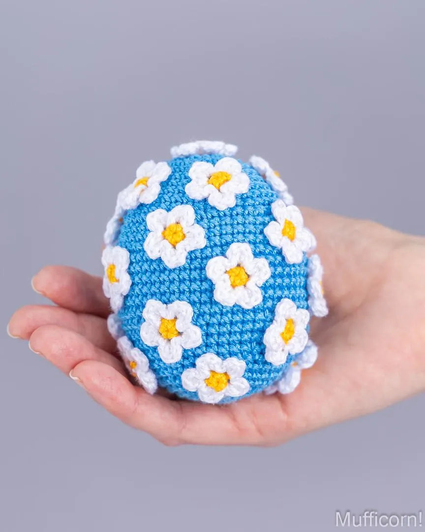 A crocheted blue egg decorated with white daisy flowers with yellow centers, held in a person's hand against a solid gray background.