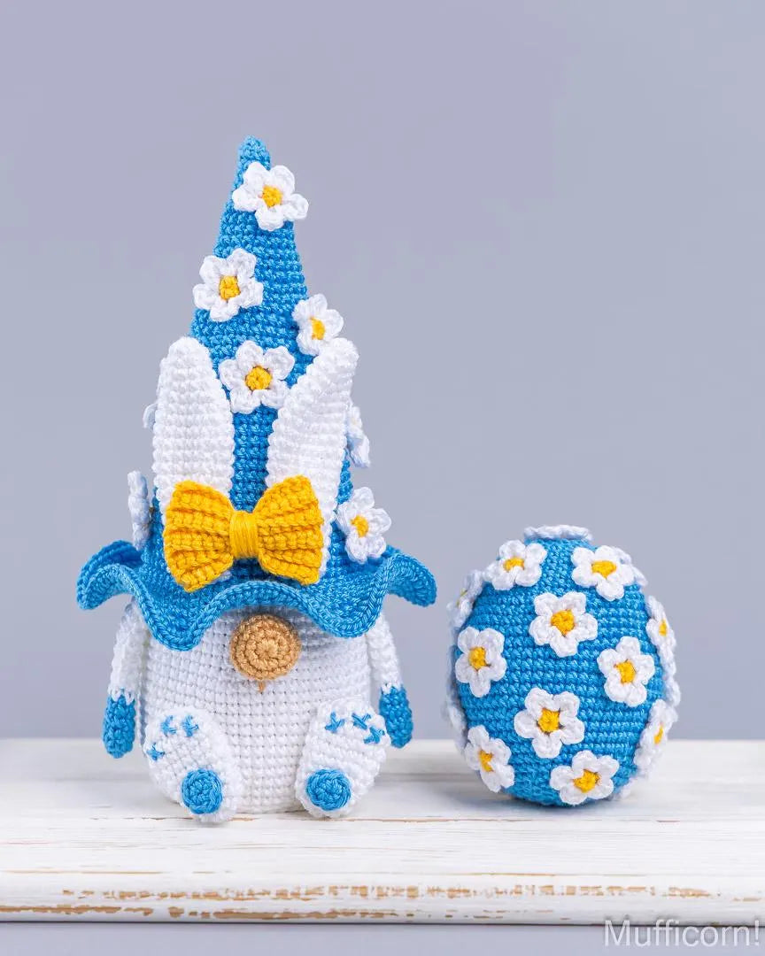 Crocheted Easter-themed decoration set featuring a blue and white bunny gnomem with a yellow bow, a matching blue and white flower-covered egg, and a blue flower hat, displayed on a white wooden surface against a gray background.