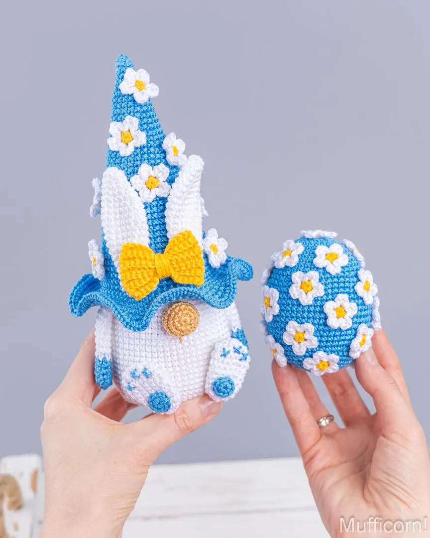 A crocheted Easter gnome figurine and matching egg, both decorated with blue and white daisy patterns and a yellow bow, held in two hands against a plain gray background.