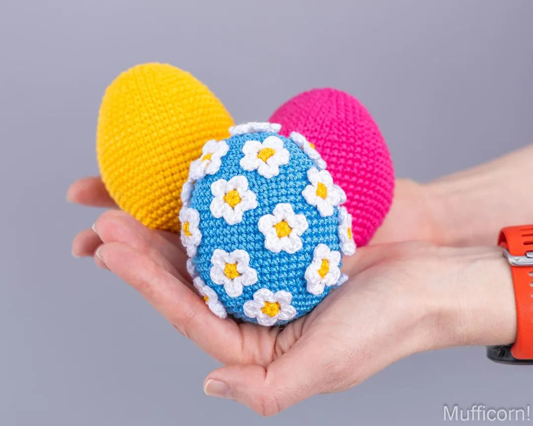 Three crocheted Easter eggs in yellow, pink, and blue with white daisy patterns, held in a person's hand against a solid gray background.