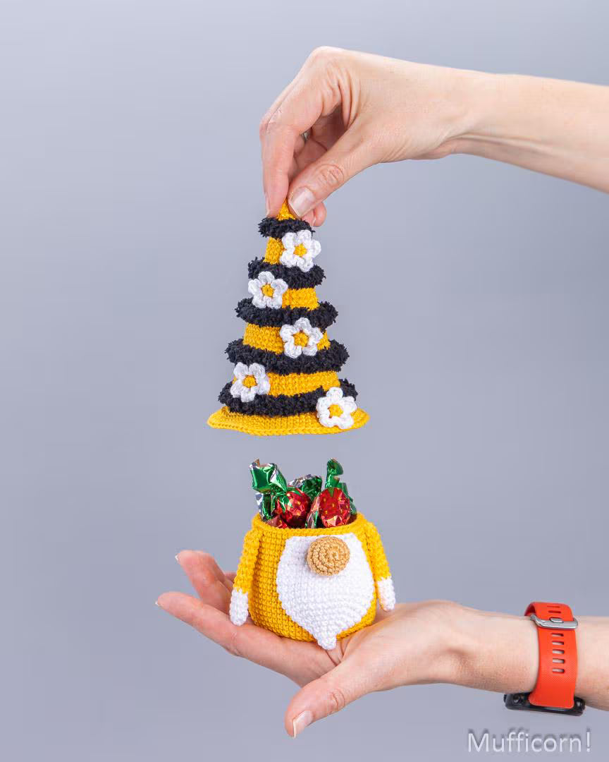 A person's hands holding a small crocheted muffin tin-shaped container on one hand and lifting a matching striped muffin topper on the other, against a plain gray background. The container is yellow and white with a round brown crumb crust, and holds small red and green candy-like decorations.
