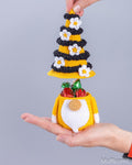 A small crocheted gnomehorn ornament with a yellow and black striped gnomehorn hat and a yellow-and-white gnomehorn body, shown being held in a person's hand against a plain gray background. The ornament is open to reveal wrapped chocolates inside, and the brand name Muffcorn is visible in the bottom right corner.